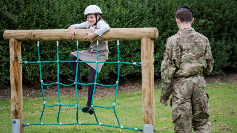 A girl climbing a green rope wall. A person in camouflage watches with their back to the camera. 